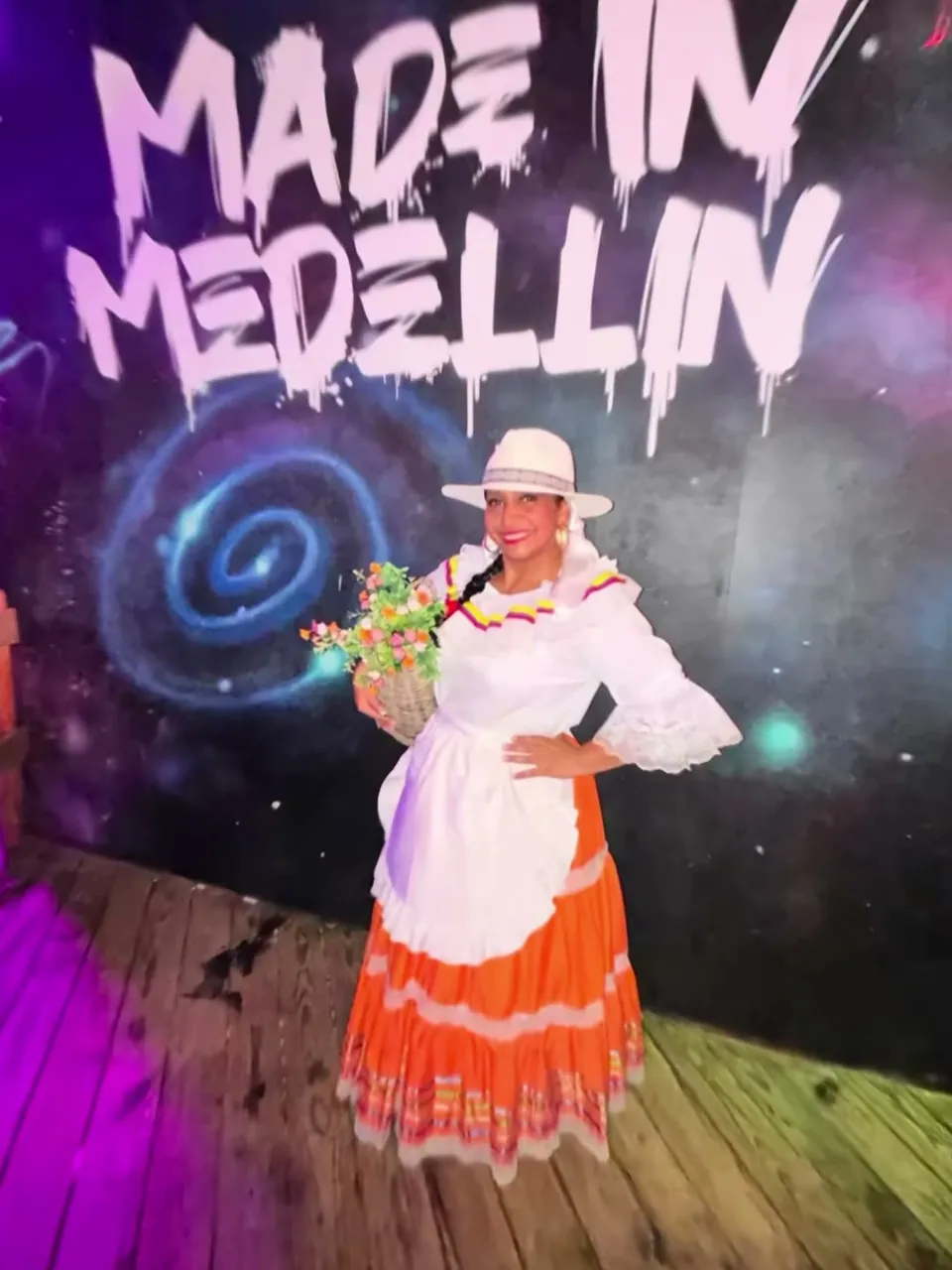 Bailarina con sombrero blanco, blusa blanca y falda naranja sosteniendo flores frente al mural de Made in Medellín