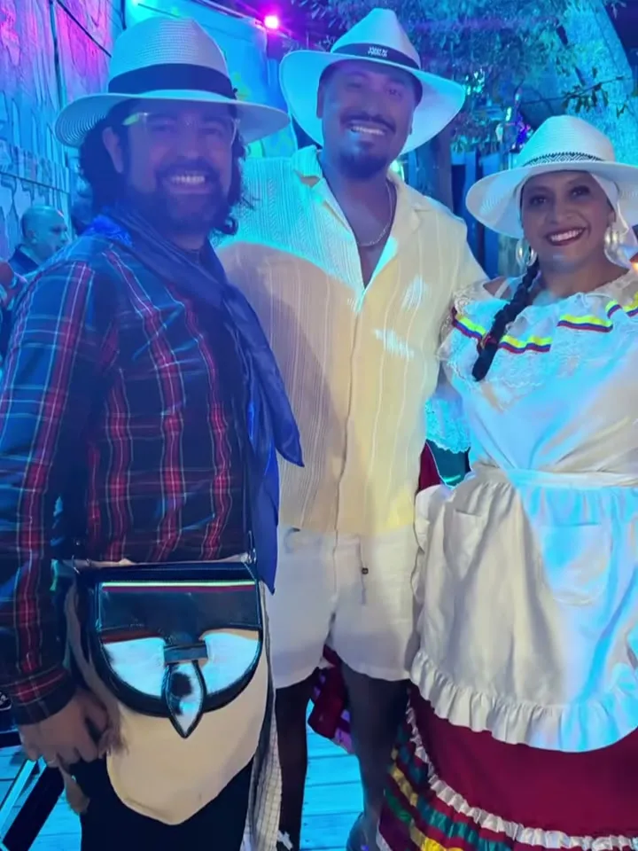 Tres personas con sombreros blancos sonriendo bajo luces azules; uno lleva ruana y carriel, otra traje típico con delantal