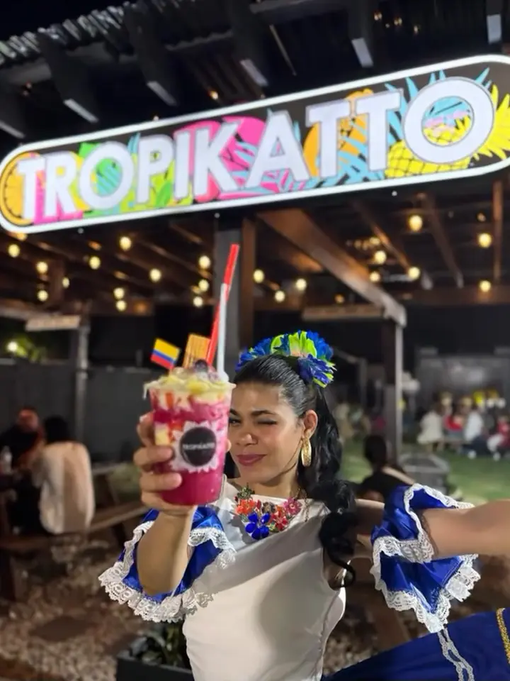 Bailarina con traje azul y blanco brindando con una bebida frente al letrero de Tropikatto